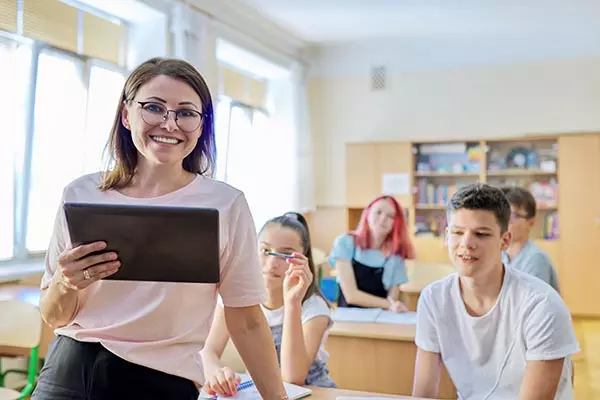 Portrait of female teacher at school at lesson, smiling middle-aged woman in glasses with digital tablet looking at camera, classroom with teenage students at desk background